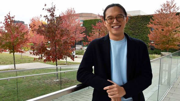 Eugene Ting wearing a blue top and a black blazer posing in front of mutiple maple trees on the York University campus