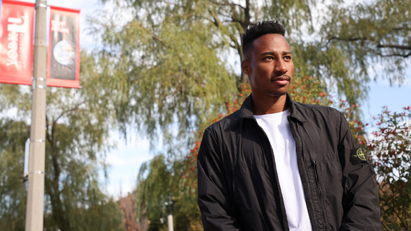 Mark-Anthony Kaye wearing a white t-shirt and black bomber jacket with York University banners in the background on the Keele Campus