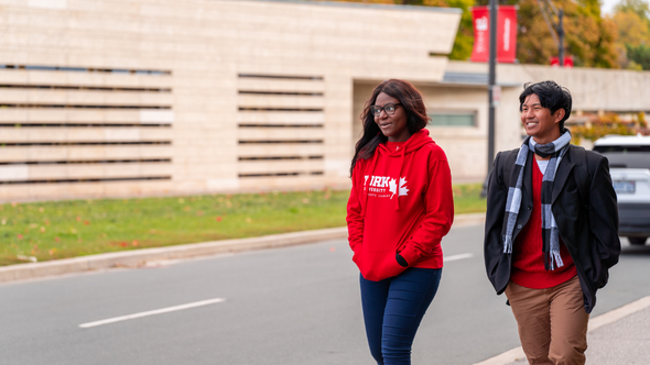 Two students walking on the York University Keele Campus, with the one in front wearing a red York U hoodie