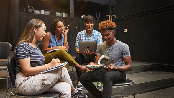 Group of diverse students sitting in dark theatre room with lighting over them while reviewing a book together