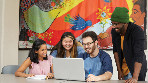 A group of diverse students looks at a laptop while smiling.
