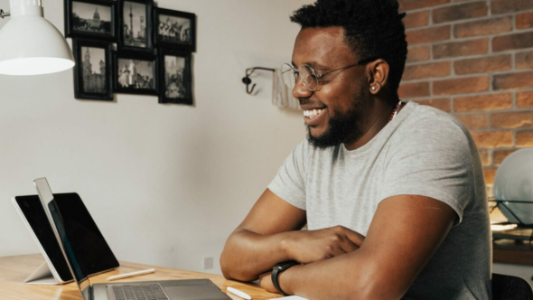 A Black man smiles while sitting on a desk and looking into a computer screen