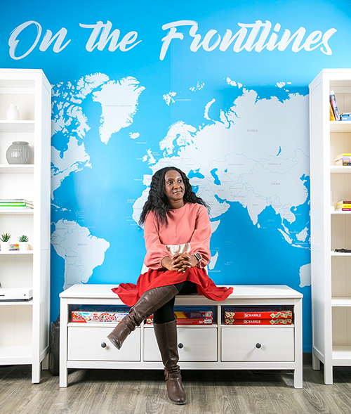 Stachen Frederick sits on a bench in front of a blue and white mural of a map of the world with the words "On The Frontlines" at the top of the wall