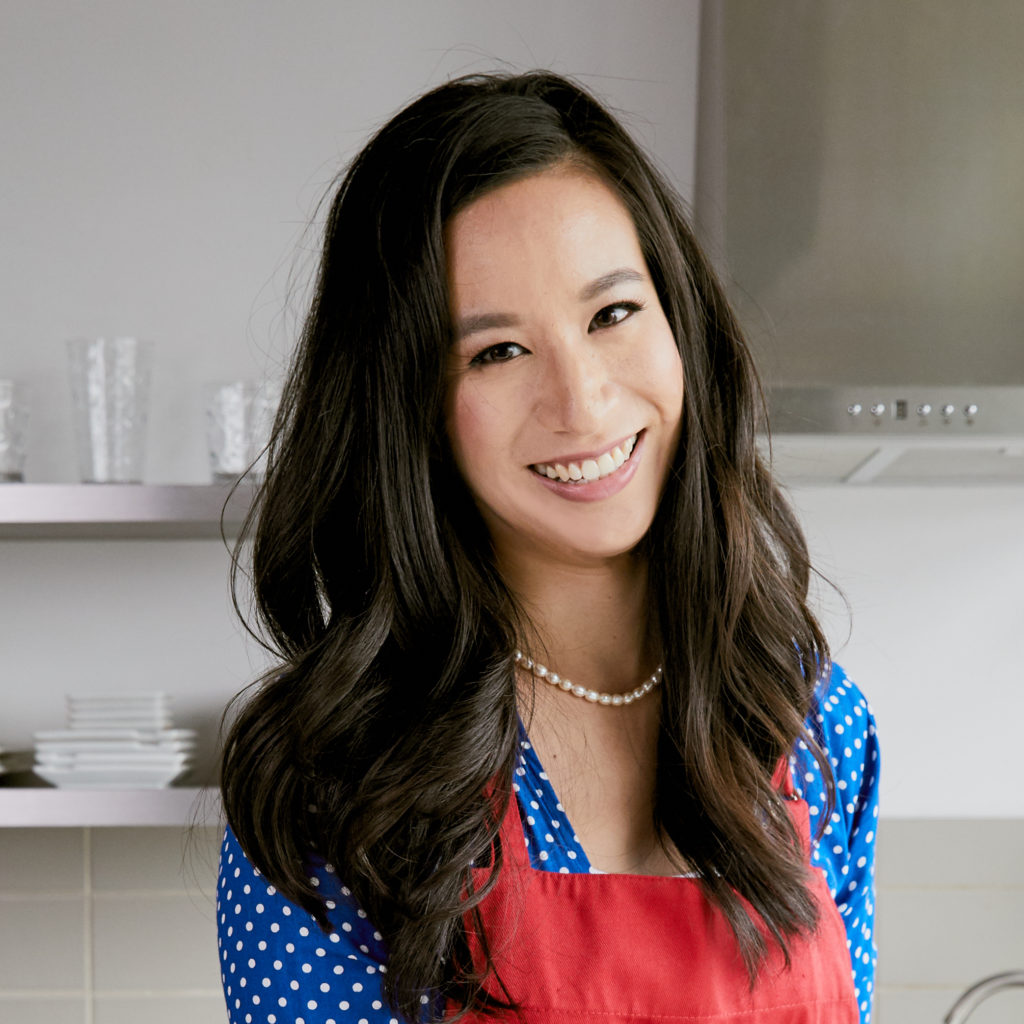 Michelle Jaelin stands in a kitchen smiling at the camera. She is wearing a red apron over a blue and white polkadot dress.