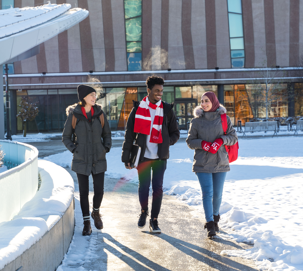Three students walk past the York University subway station. They are dressed for winter, there is snow on the ground and you can see their breath in the air.