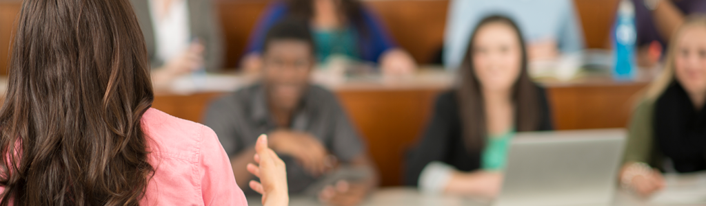 woman in pink blouse speaks at the front of a lecture hall of students