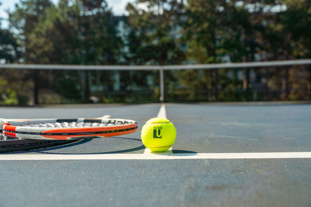 Tennis racquet and yellow tennis ball with York U logo on a tennis court