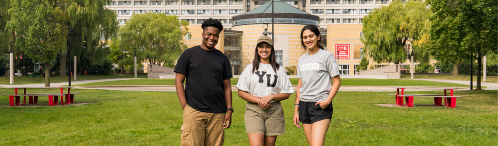 3 diverse students stand in front of Vari Hall in the grass area.
