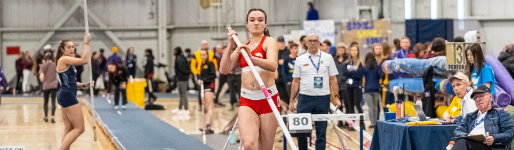 A young woman in track and field clothing runs down a lane while holding a pole preparing for a leap.