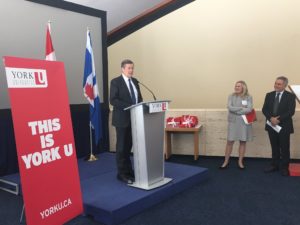 A man in formal clothing speaks at a podium stationed in front of the Canadian and Ontario flags. There is a red York banner that reads "THIS IS YORK U" to his right, and a man and a woman standing at his left side.
