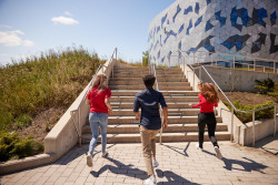 Three students are running towards a flight of stairs in front of a glass building.