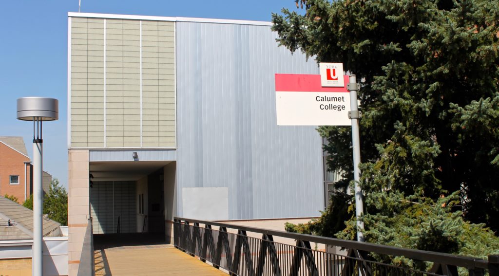 A beige and grey building stands behind a wooden bridge with black railings. A sign in front of the building reads "Calumet College".