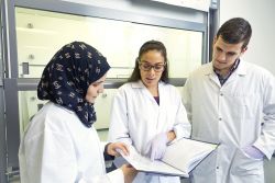 Three students in lab coats are looking through a notebook.