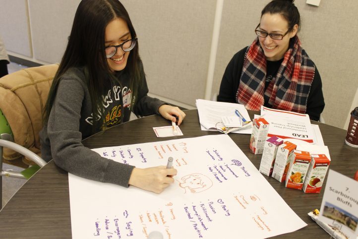 Two students are sitting at a table together. One student is writing on a piece of paper.