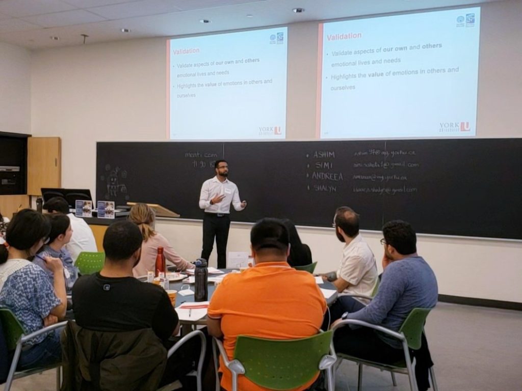 A presenter standing next to the lecture board. Students are grouped together and listening to the presenting.