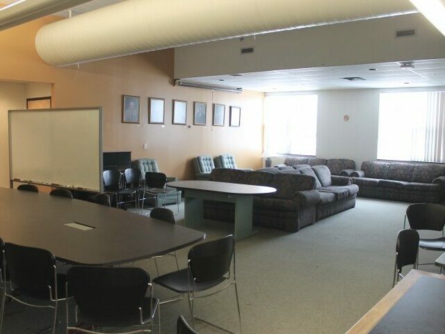 A room with a grey carpet and light-brown walls. Grey tables are organized around the room along with chairs and couches.