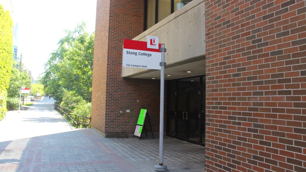 A dark red and black brick building with grey cement accents. A sign in front of the building reads "Stong College". 