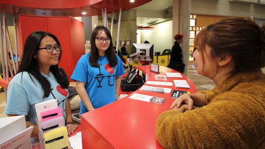 Two women wearing I love YU t-shirts stand behind desk while helping a student in Scott Library on York University Keele Campus