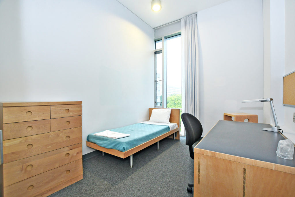 A single room in a Pond Road Residence suite showing bed, dresser, desk, lamp, window.