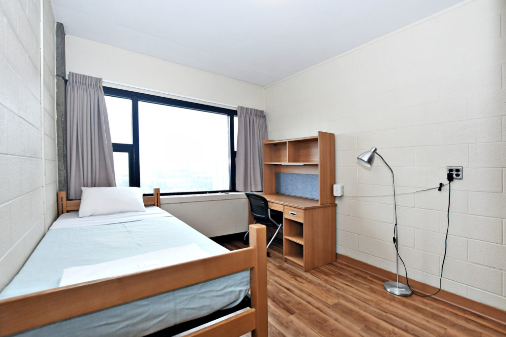 A single dorm room showing bed, desk, lamp, window.