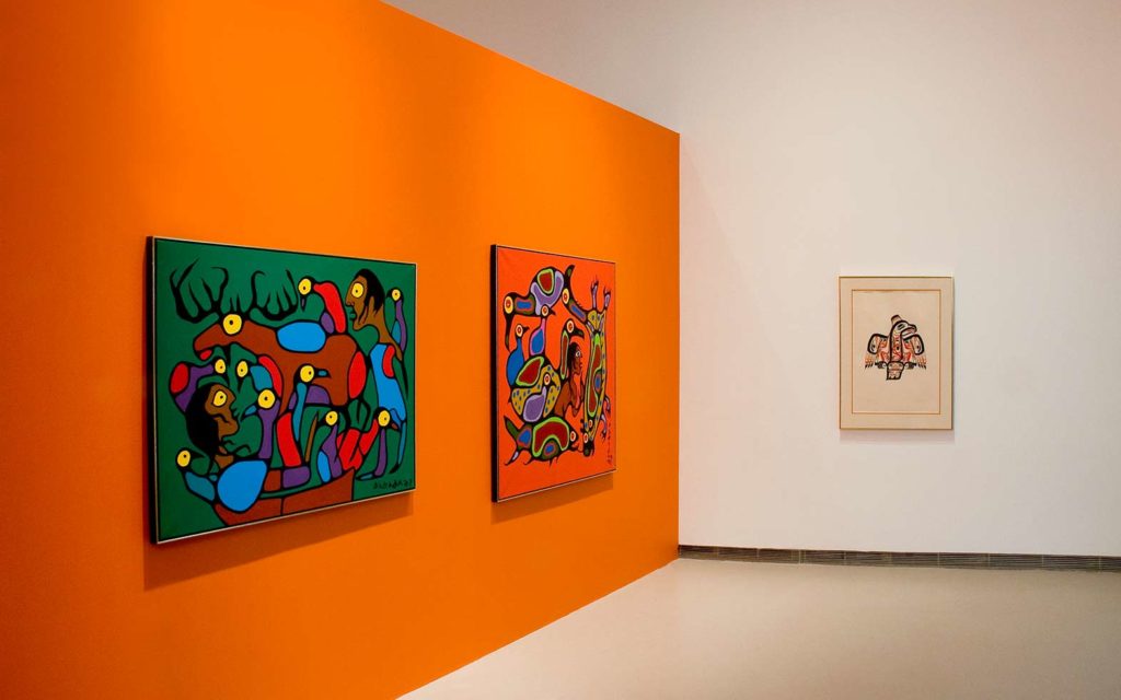 Art pieces hang on an orange wall in a gallery.