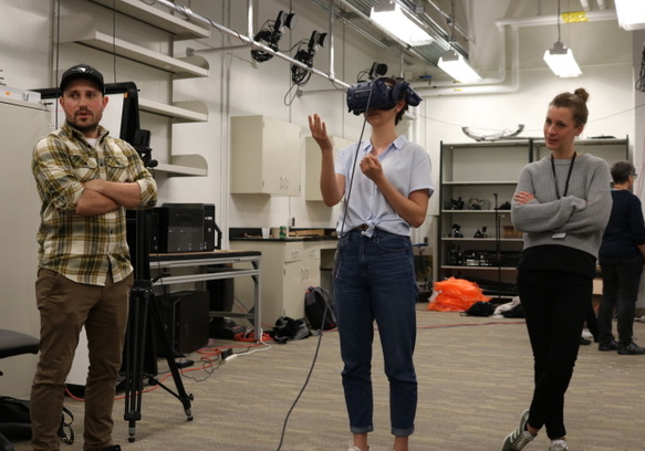 Person standing and wearing VR headset. Two people stand beside with their arms crossed.