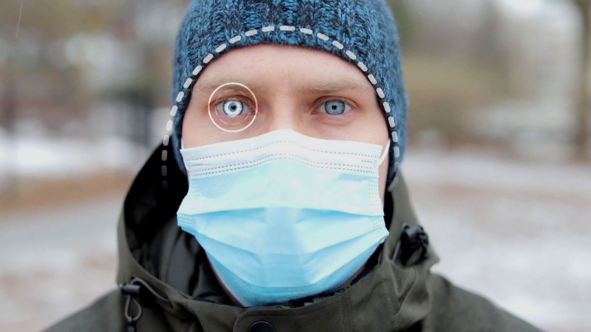Face of a person wearing a blue face mask, jacket, and hat looking straight at the camera. A recognition lens is placed on their eye.