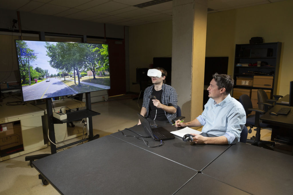 Person wearing a VR headset at a table with someone else who is looking at TV displaying the VR images.