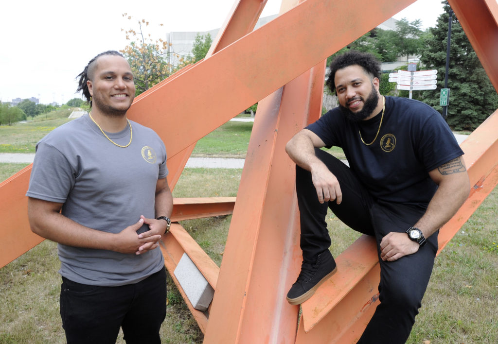 Jonatan Fuentes (left) and his brother, Ryan Fuentes (right) outside in a park posing in front of a large metal abstract art structure.