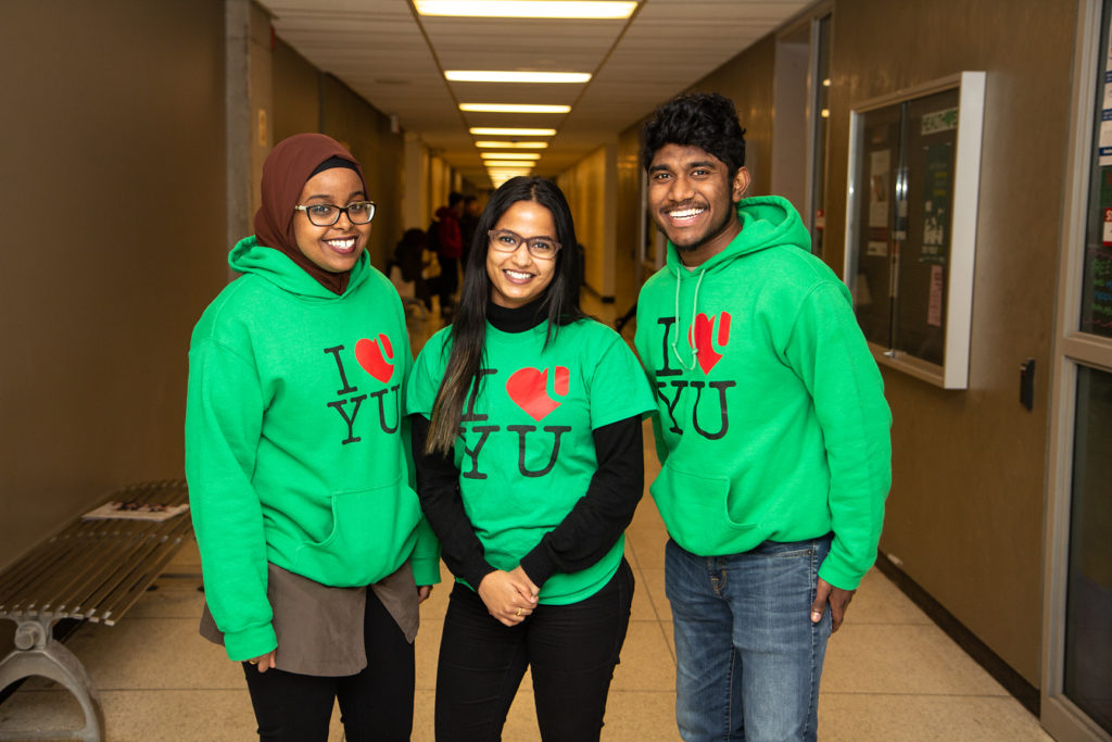 image of 3 smiling students wearing green 'I love YU' sweatshirts. Pictured from left to right: female student wearing a hijab; female student; male student)