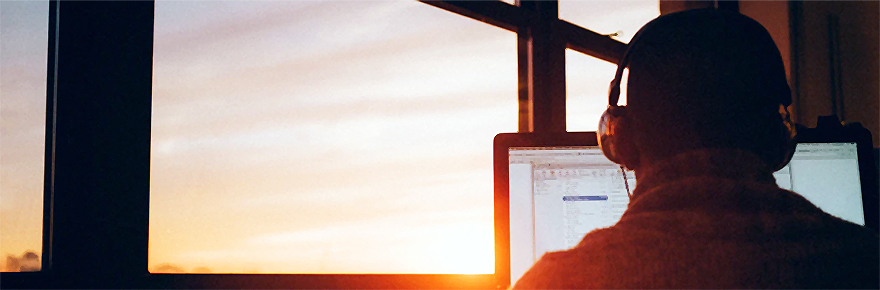 Man wearing a headset doing a seminar in front of a computer screen. Man is sitting facing a window that is displaying a sunset outside.