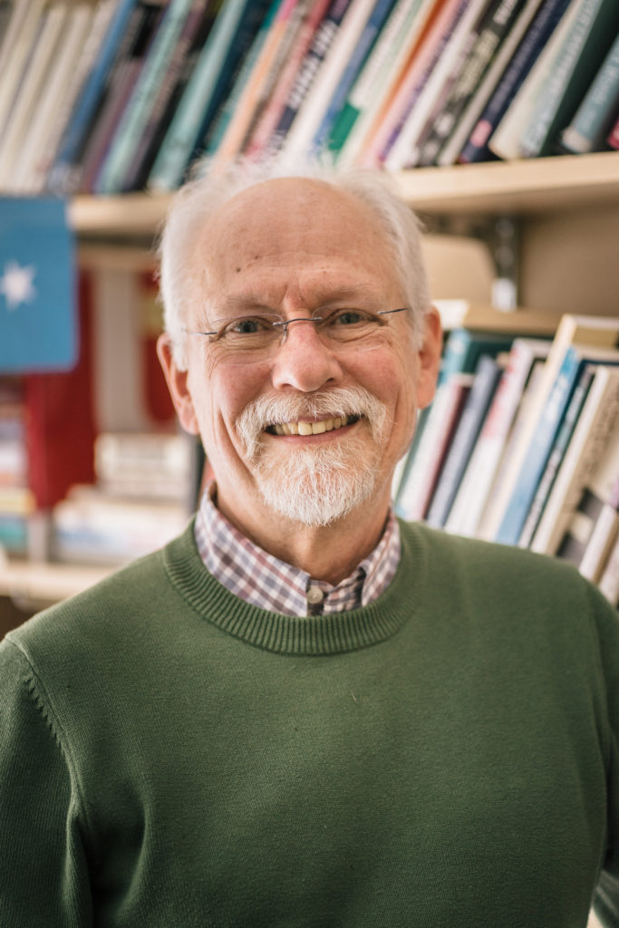 headshot of professor Don Dippo wearing a hunter green sweater and standing in front of a bookshelf in his office