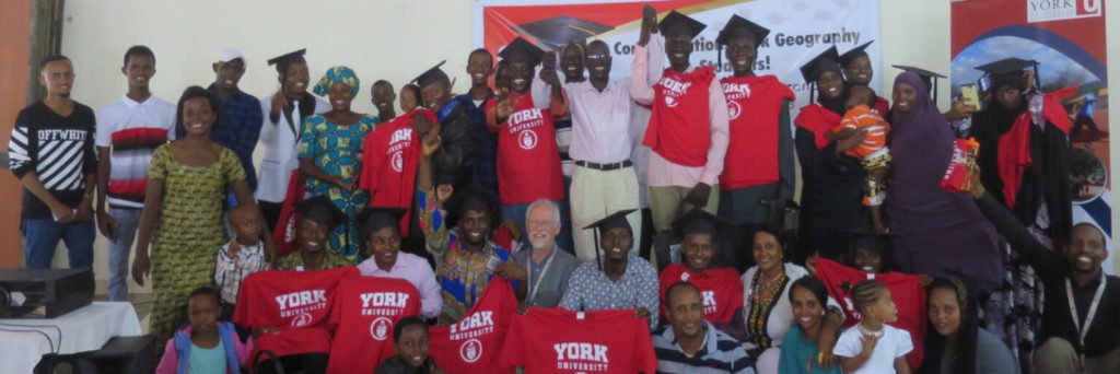 students and their families who are part of the BHER program posing at a graduation ceremony at the Education Centre in the Dadaab camp. Some of the students are wearing/holding red York University t-shirts.