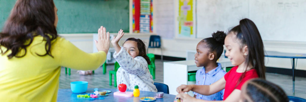 female teacher in an elementary classroom giving a high five to a young female student who is sitting beside 2 other young female students
