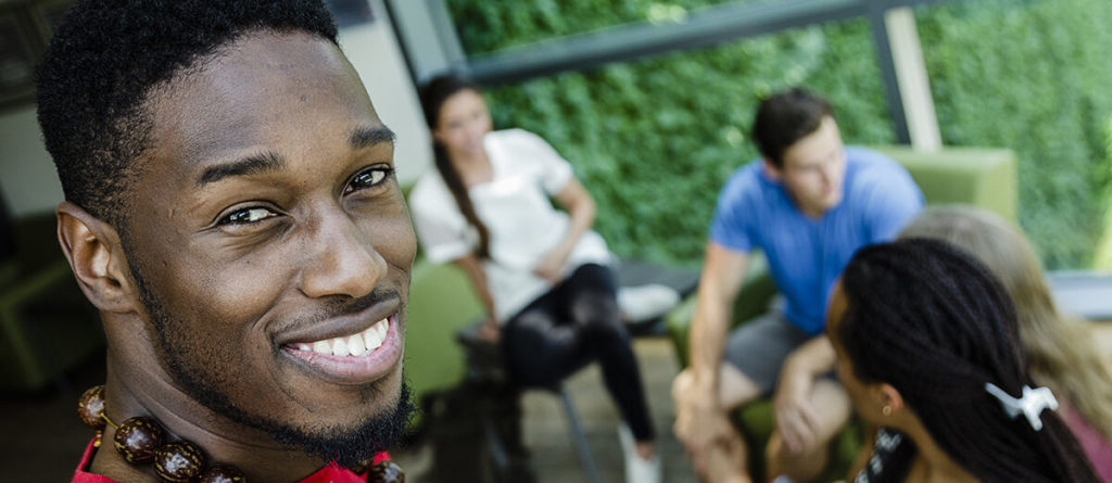 Young black man looking back and smiling at camera while sitting in a lounge with friends