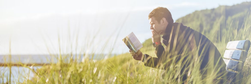 Man sitting outside on a bench in a field by a lake reading a book