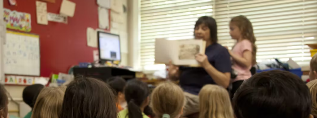 White middle-aged female teacher sitting in front of a classroom reading a book to her students