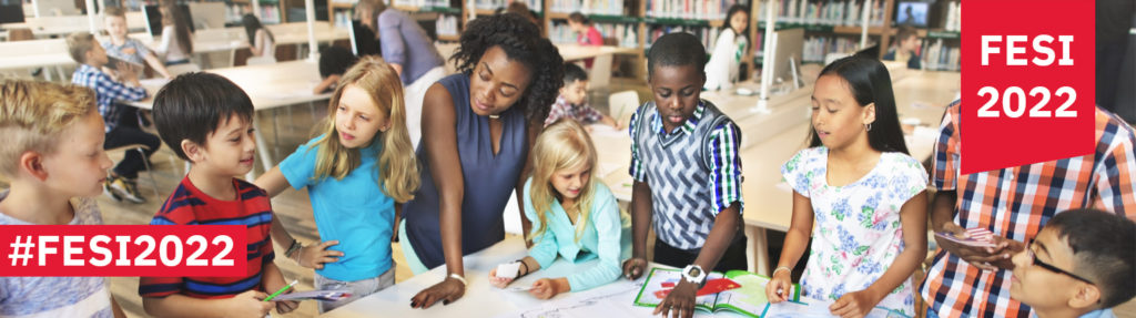 image of young female black teacher standing around a table with a group of diverse elementary students working on a project