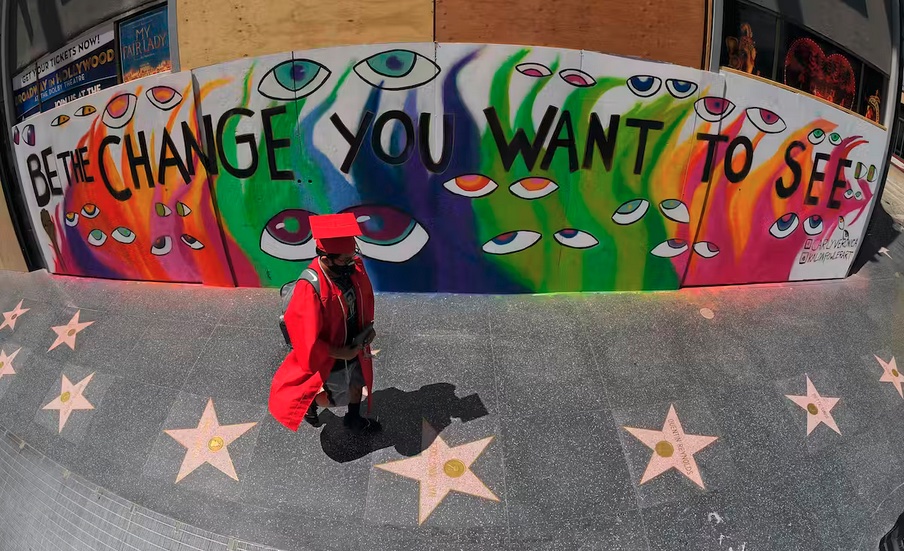 A student dressed in graduation gown and hat walks in front of a colourful mural that reads "Be the change you want to see"