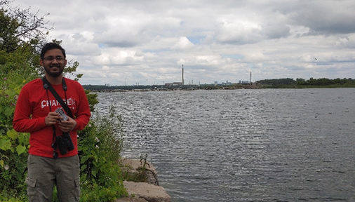 Meetkumar - a SEM Students in EUC-  at Tommy Thompson Park holding a trail camera with a cormorant colony in the background.