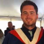 A young man with facial hair smiles while wearing a graduation gown and hood.