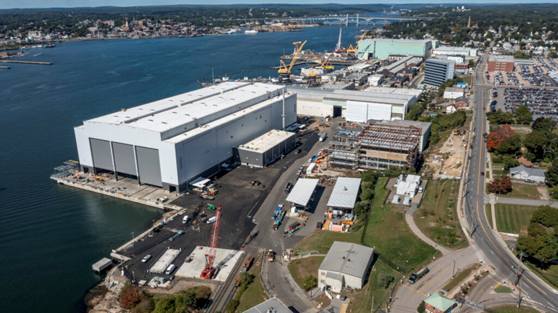 An aerial view of a large industrial shipyard along a waterfront, featuring massive white warehouses and construction cranes.