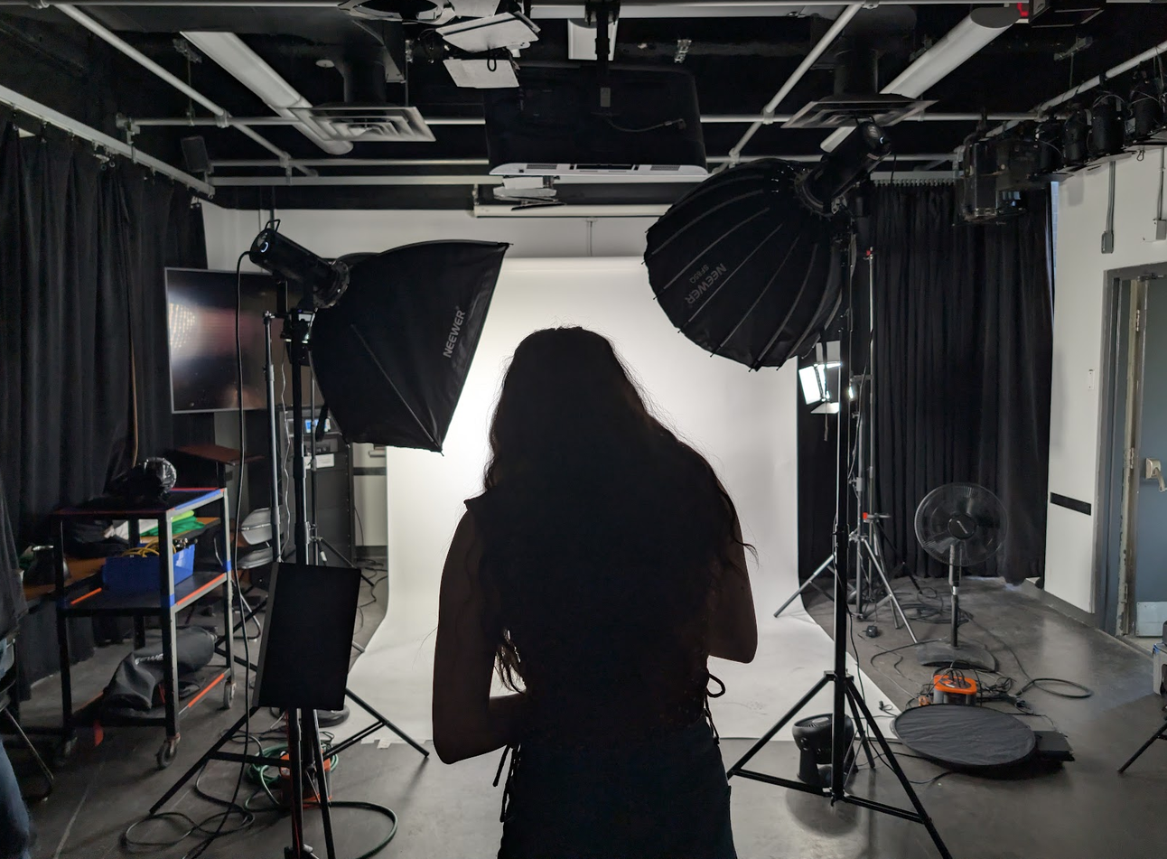 Behind-the-scenes view of a photography studio, with a person standing in front of a white backdrop surrounded by professional studio lights and equipment.