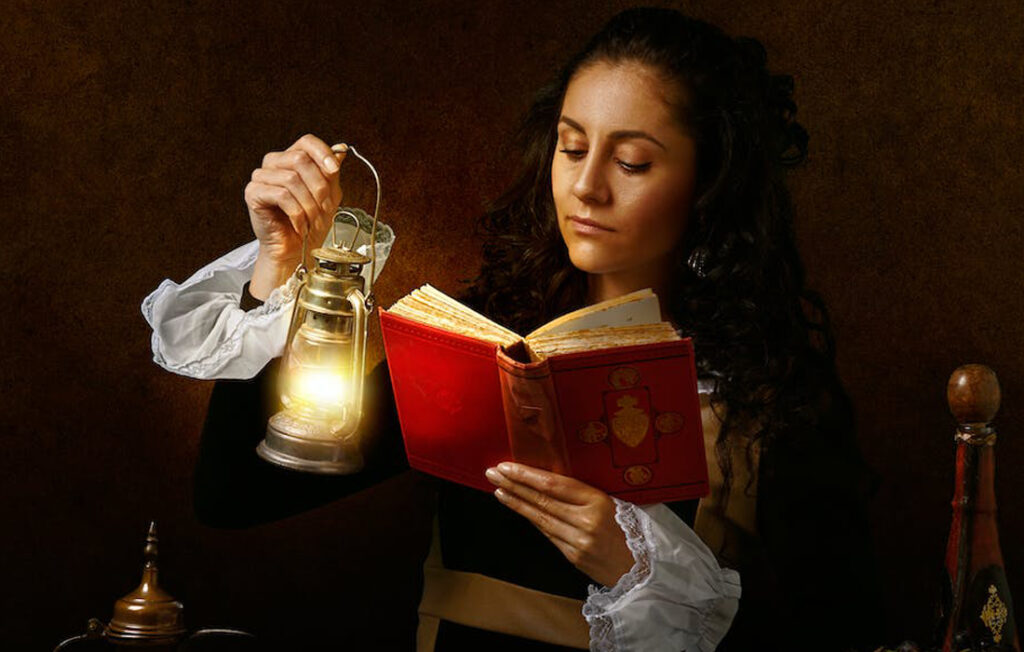 woman in old outfit reading book with oil lamp