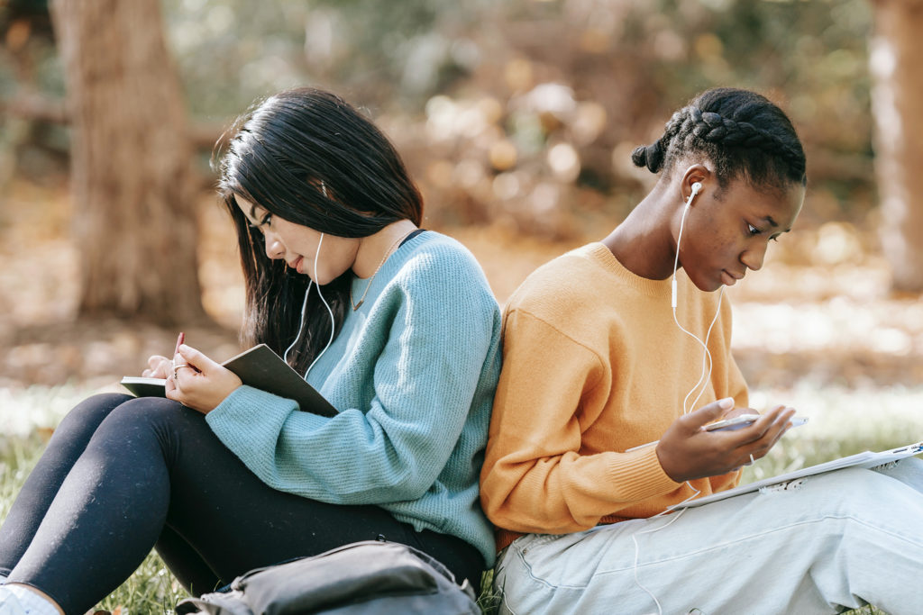 image of two young women with ear buds outside writing in notebooks