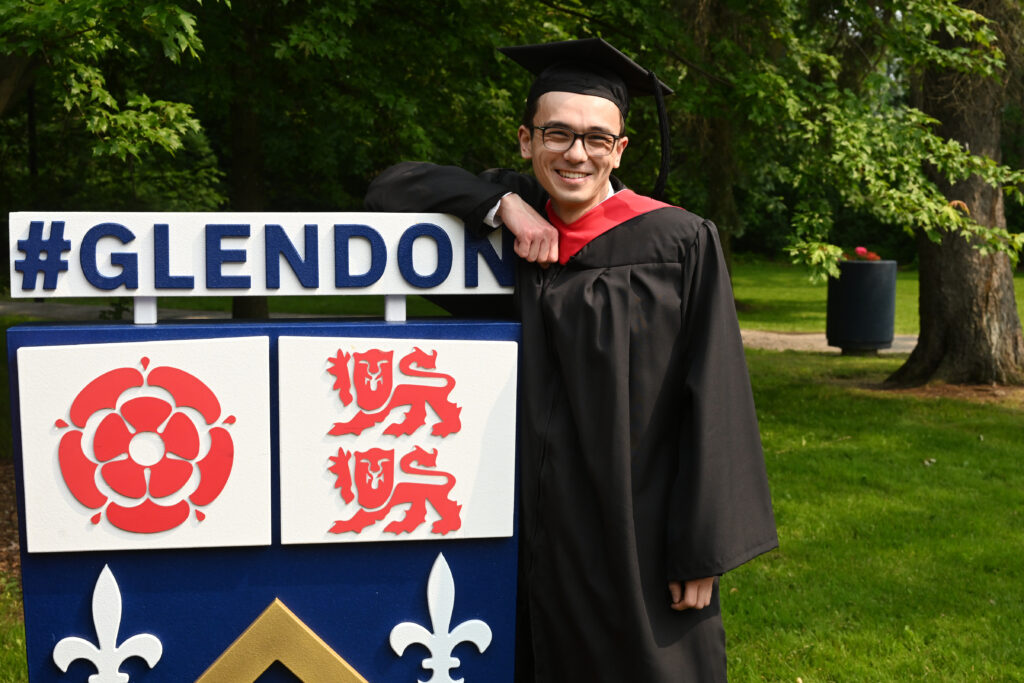 A university graduate standing and smiling is wearing a black cap and gown stands with one arm on top of a Glendon College sign.