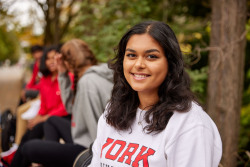 One YorkU student is in the foreground smiling and wearing a gray York sweatshirt and there are 3 students in the background sitting on a bench.