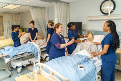 Six YorkU students in nursing uniforms are standing at hospital beds and one patient artificial model is in a bed in a hospital setting.