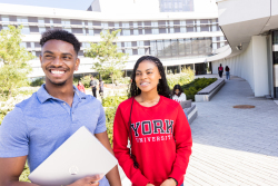 Two YorkU students are standing outside and smiling while looking into the distance. One student is wearing a read York sweatshirt and one student is wearing a blue shirt and holding a white notebook.