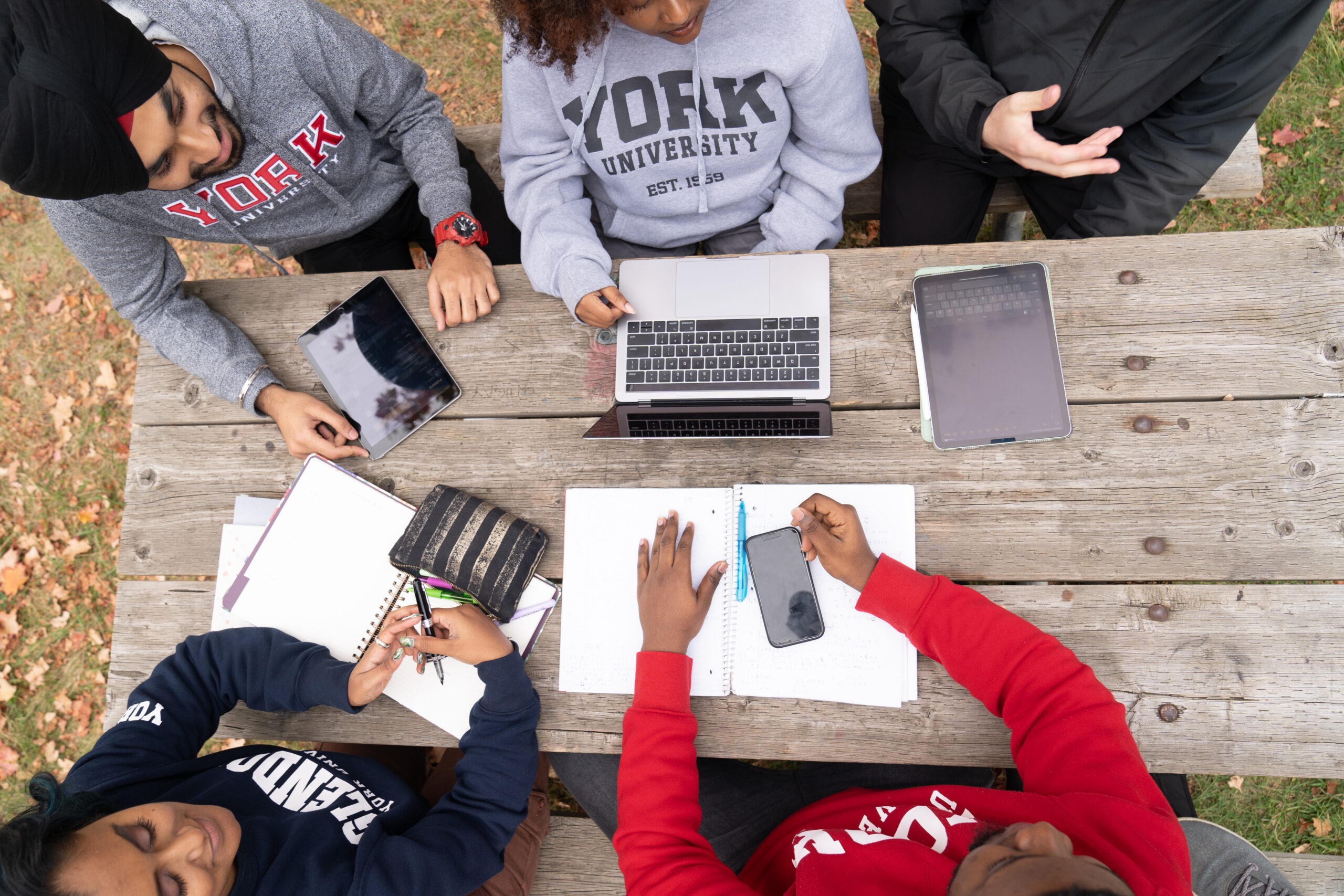 A group of 5 students sitting outdoors at a picnic table facing each other and there are 2 tablets, 1 laptop and 1 smart phone with 2 paper notebooks and pens on the table. The students are dress in red, blue, light gray and dark gray York sweatshirts.